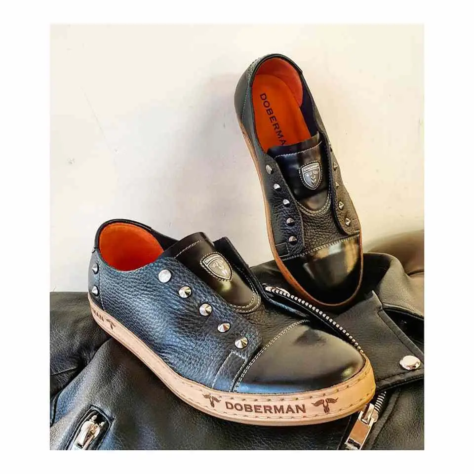 A pair of  black leather slip-on sneakers for men with stud details, a shiny toecap, and a light-colored sole.