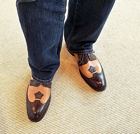 A close-up, three-quarter view of a single men's two-tone Goodyear Welted brogue oxford shoe. The shoe is dark brown on the toe, heel, and lacing, and tan on the sides and top, with decorative perforations outlining the sections.